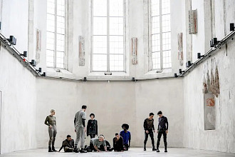 Zehn Tänzer:innen befinden sich in einem hellen, weißen Raum mit hohen Decken, der an eine alte Kirche erinnert. Auf der linken Seite beobachtet eine stehende Person fünf weitere, die auf dem Boden sitzen und knien. Diese blicken auf vier weitere Personen. Zwei stehen zu ihrer Rechten, eine steht vor der Gruppe mit dem Rücken zur Kamera und eine weitere steht im Hintergrund und blickt nach vorne.