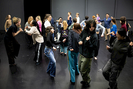 Ungefähr zwanzig Personen tanzen. Im Vordergrund bewegen mehrere Personen ihre Arme in gebeugter Haltung und neigen ihren Oberkörper leicht zur Seite. Im Hintergrund gehen und drehen sich andere Teilnehmer.