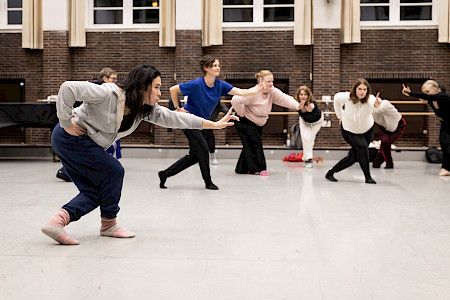 Acht Personen stehen in einem großen Tanzstudio. Im Vordergrund beugt eine Person die Beine und streckt einen Arm nach vorne, während die anderen im Hintergrund dieselbe Haltung einnehmen.