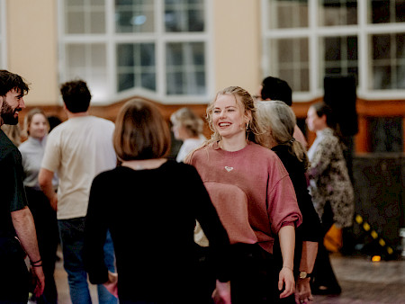 Eine Gruppe von Personen tanzt miteinander und bewegt sich in verschiedene Richtungen. In der Mitte lächelt eine Person, während sie sich auf eine andere zubewegt. Im Hintergrund kreuzen sich mehrere Personen und interagieren miteinander.
