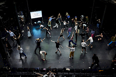 Auf einer Theaterbühne tanzt eine Gruppe von ungefähr dreißig Personen über die Bühne. Einige heben ihre Arme, andere beugen sich oder lehnen sich zur Seite; alle machen weite, schwungvolle Armbewegungen. Im Hintergrund zeigt eine Leinwand den Text “Welcome to the Tanztriennale.”