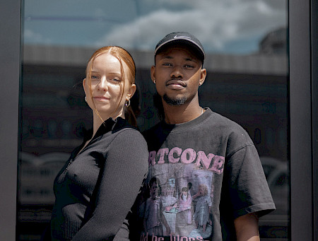 Dunkler Hintergrund. In der Mitte stehen zwei Personen Rücken an Rücken und blicken in die Kamera. Eine trägt ein schwarzes T-Shirt und eine Kappe; die andere ein schwarzes Langarmoberteil und hat die Haare zurückgebunden. Beide lächeln leicht.