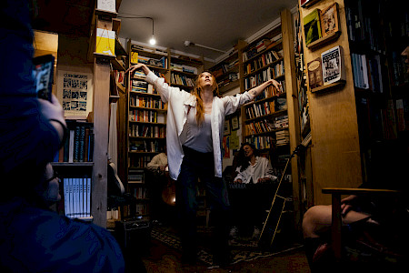 In einer Buchhandlung steht eine Person mit nach oben erhobenen Armen und gebeugten Händen. Sie trägt ein offenes weißes Hemd über einem hellen Oberteil und eine dunkle Hose. Im Hintergrund sitzt eine weitere Person und spielt Keyboard. Weitere Personen schauen vom Rand des Bildes zu.