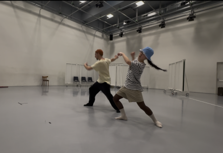 In einem Tanzstudio mit weißen Wänden und grauem Boden tanzen zwei Personen. Beide stehen mit gespreizten Beinen, ein Bein vorne gebeugt und das andere nach hinten gestreckt. Der Oberkörper ist nach hinten geneigt und die Arme sind seitlich geöffnet.