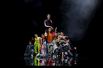 In der Mitte steht eine Person und trägt eine andere auf ihren Schultern. Hinter ihnen schaut eine Gruppe von Personen zu, einige kniend, andere sitzend oder mit ausgestreckten Armen. Sie tragen farbenfrohe, auffällige Kleidung. Die Beleuchtung hebt die Gruppe vor dem schwarzen Hintergrund hervor.