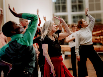 Eine Gruppe von Personen nimmt an einem Tanzkurs teil. Vor ihnen hebt eine Person einen Arm und neigt den Kopf sanft nach hinten. Um sie herum wiederholen andere die gleiche Bewegung mit erhobenen Armen.