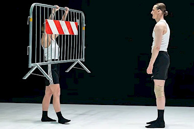 Auf einer Bühne mit weißem Boden und schwarzen Wänden stehen sich zwei Tänzer:innen gegenüber. Beide tragen schwarze Strümpfe, schwarze Shorts und weiße, ärmellose Hemden. Die performende Person auf der linken Seite hält eine Metallbarriere mit einem rot-weißen Streifen, der daran hängt. Ihr gegenüber, auf der rechten Seite des Bildes, steht die zweite performende Person und sieht sie aufmerksam an.