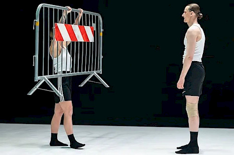 Auf einer Bühne mit weißem Boden und schwarzen Wänden stehen sich zwei Tänzer:innen gegenüber. Beide tragen schwarze Strümpfe, schwarze Shorts und weiße, ärmellose Hemden. Die performende Person auf der linken Seite hält eine Metallbarriere mit einem rot-weißen Streifen, der daran hängt. Ihr gegenüber, auf der rechten Seite des Bildes, steht die zweite performende Person und sieht sie aufmerksam an.