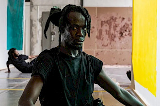 Eine Person mit hochgebundenem Haar sitzt seitlich auf dem Boden eines hellen, industriell wirkenden Raums, ein Bein angewinkelt, das andere nach vorn ausgestreckt, und blickt direkt in die Kamera. Neben ihr hängen große Stoffbahnen in Türkis und Gelb, im Hintergrund liegt eine weitere Person am Boden.