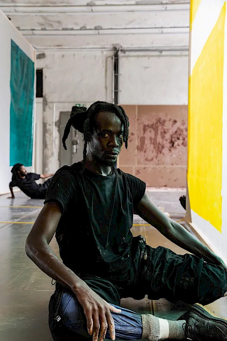 Eine Person mit hochgebundenem Haar sitzt seitlich auf dem Boden eines hellen, industriell wirkenden Raums, ein Bein angewinkelt, das andere nach vorn ausgestreckt, und blickt direkt in die Kamera. Neben ihr hängen große Stoffbahnen in Türkis und Gelb, im Hintergrund liegt eine weitere Person am Boden.