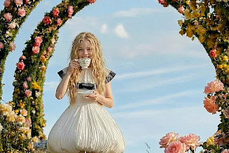 Ein Kind mit langen hellen Haaren steht auf einer Wiese unter mehreren blumenumrankten Bögen und hält eine Tasse mit Untertasse vor sich. Es trägt ein helles, ballonförmiges Kleid, umgeben von rosa, gelben und weißen Blüten.